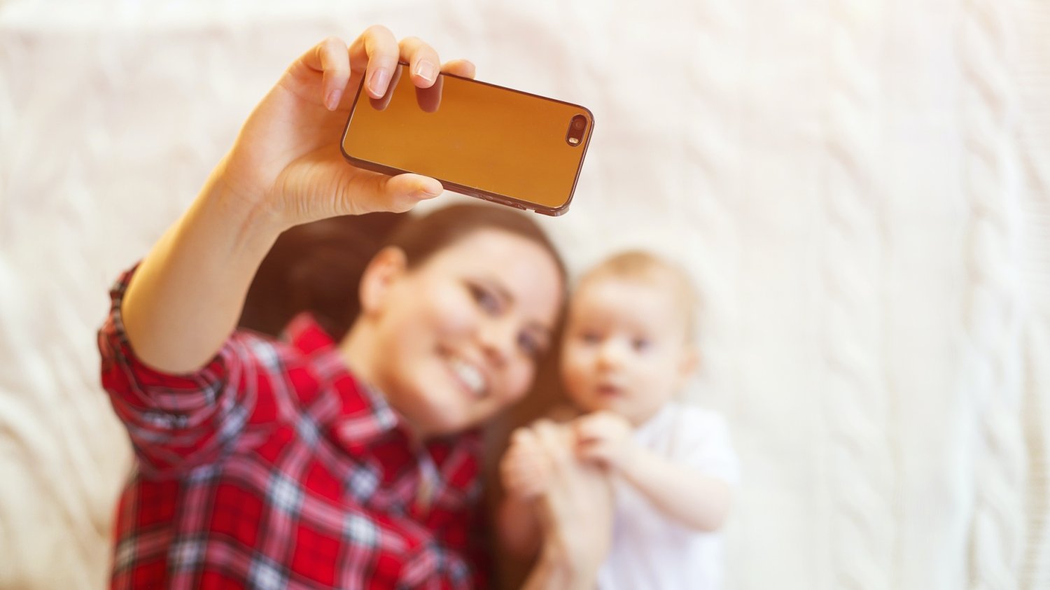 Trendy rodzicielskie, które powinniśmy pożegnać w 2026 roku Na zdjęciu mama robi sobie zdjęcie z dzieckiem (niemowlakiem leżącym na łóżku)