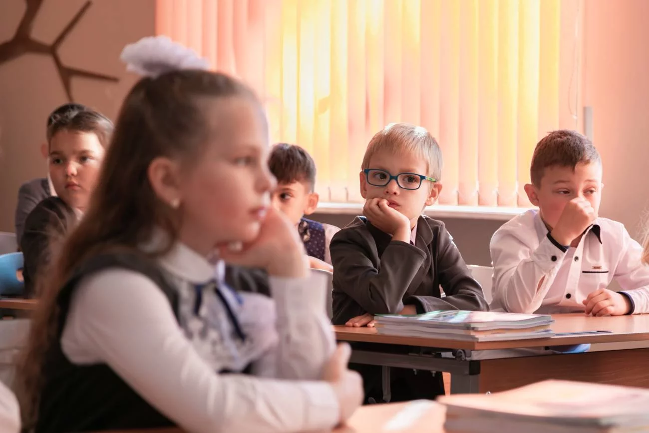 Dzieci siedzą na lekcji w ławkach i słuchają nauczyciela.