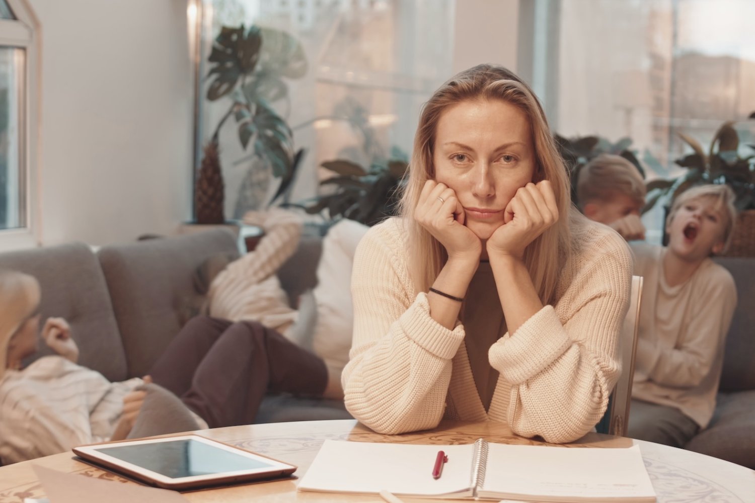 Na zdjęciu kobieta (mama) siedzi przy stole, za nią dzieci rozrabiają na kanapie.