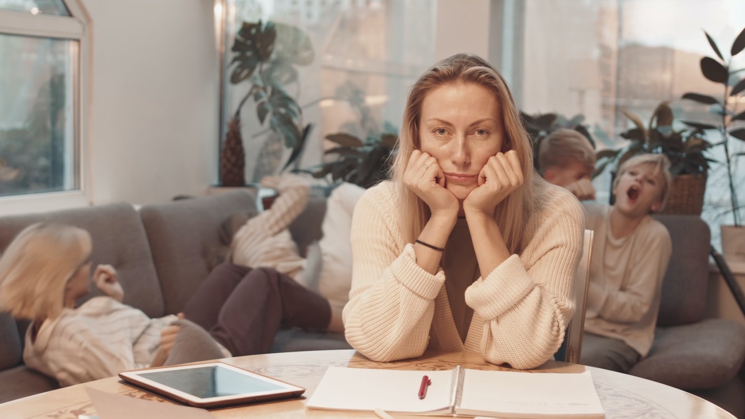 Na zdjęciu kobieta (mama) siedzi przy stole, za nią dzieci rozrabiają na kanapie.