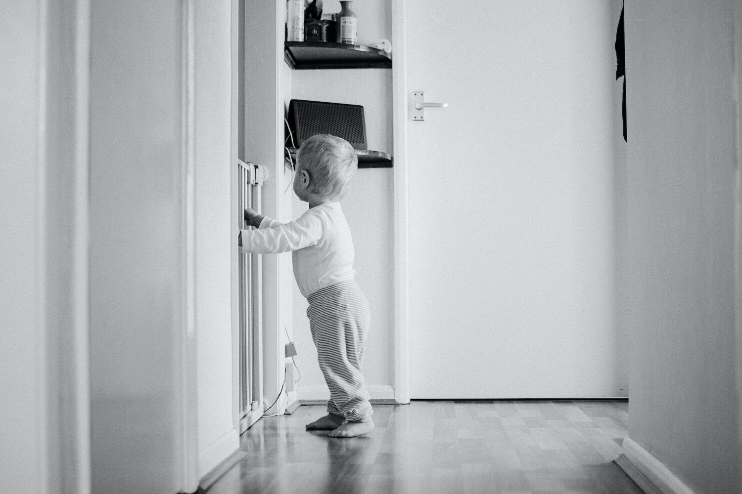 Małe dziecko stoi w mieszkaniu, opiera rączki o bramkę bezpieczeństwa.