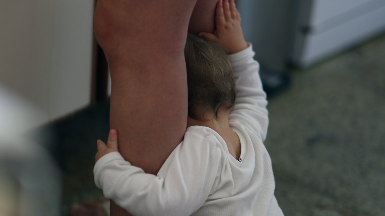 Dziecko rok po roku Na zdjęciu małe dziecko trzyma mamę, przygotowującą coś w kuchni, za nogi