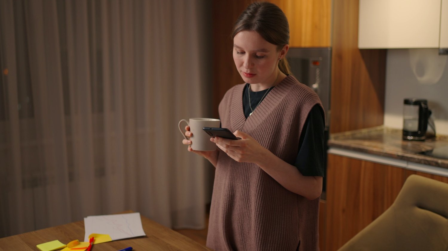 Na zdjęciu kobieta stoi w salonie i wpatruje się w telefon.
