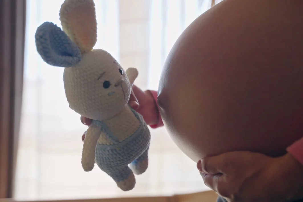 Dlaczego niektórzy mają same córki albo samych synów? Na zdjęciu widać kobiecy brzuch ciążowy. Przyszła mama trzyma króliczka z różowo-niebieskimi uszami.