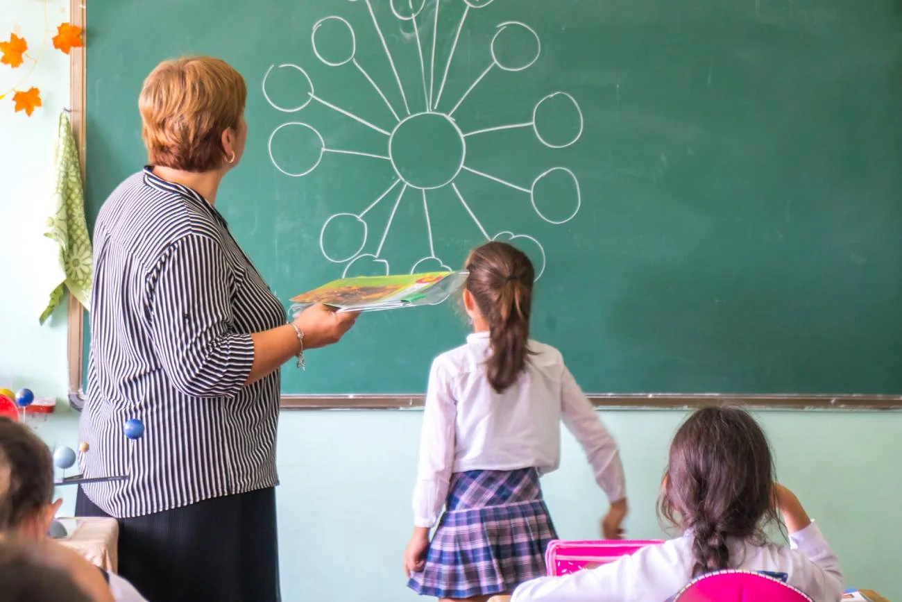Nauczycielka klasy 1 prowadzi lekcję i tłumaczy coś przy tablicy dziewczynce.