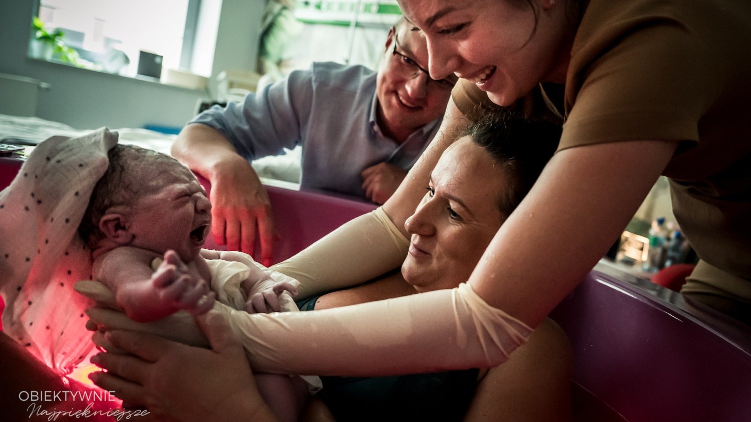 Fotografia porodowa – wywiad z fotografką Anią Wibig Na zdjęciu widać rodzącą kobietę z dzieckiem (poród odbywa się w basenie) na drugim planie jest położna i prawdopodobnie partner kobiety i ojciec dziecka