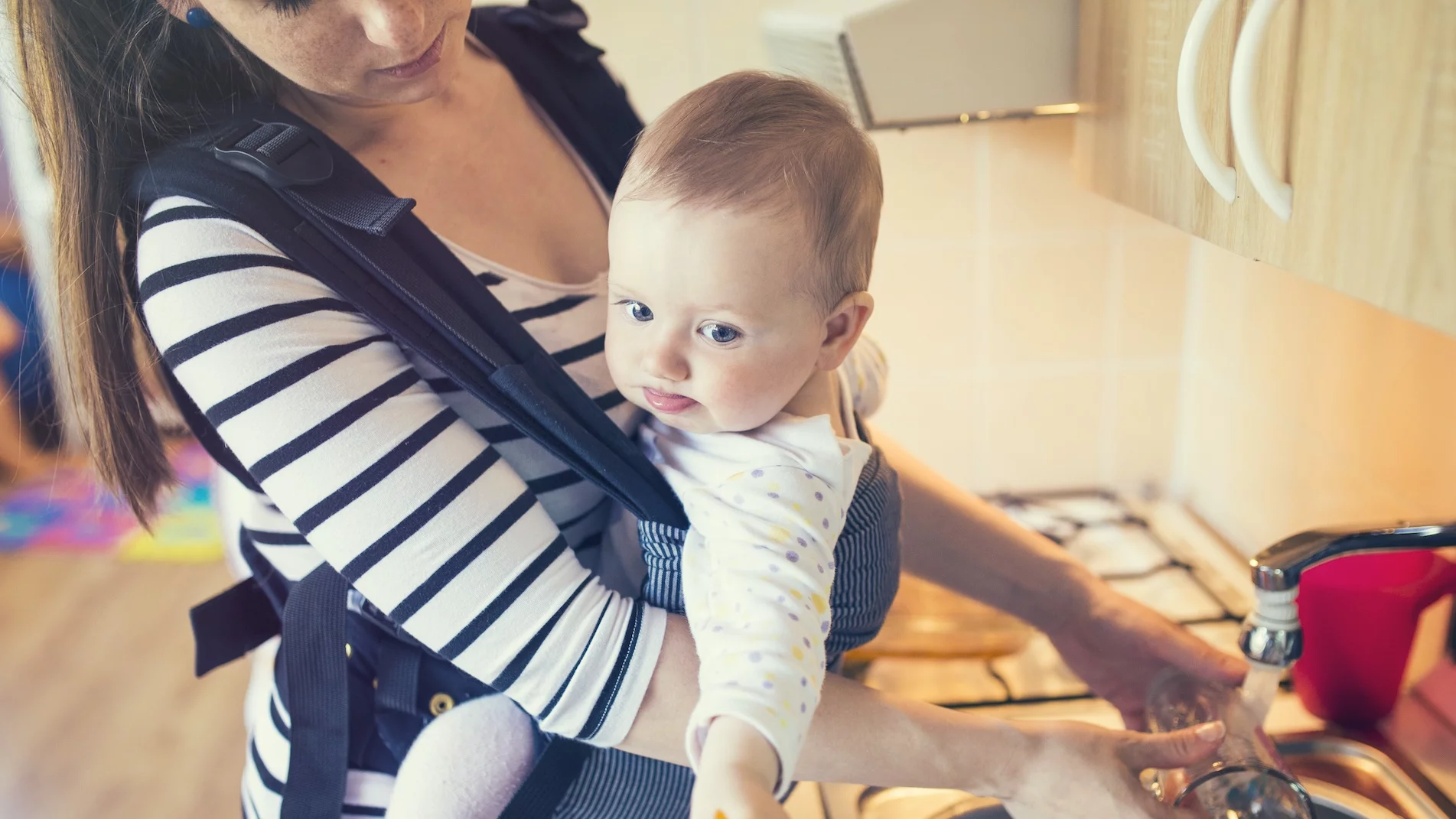 Na zdjęciu kobieta trzyma dziecko w nosidełku i zmywa naczynia.