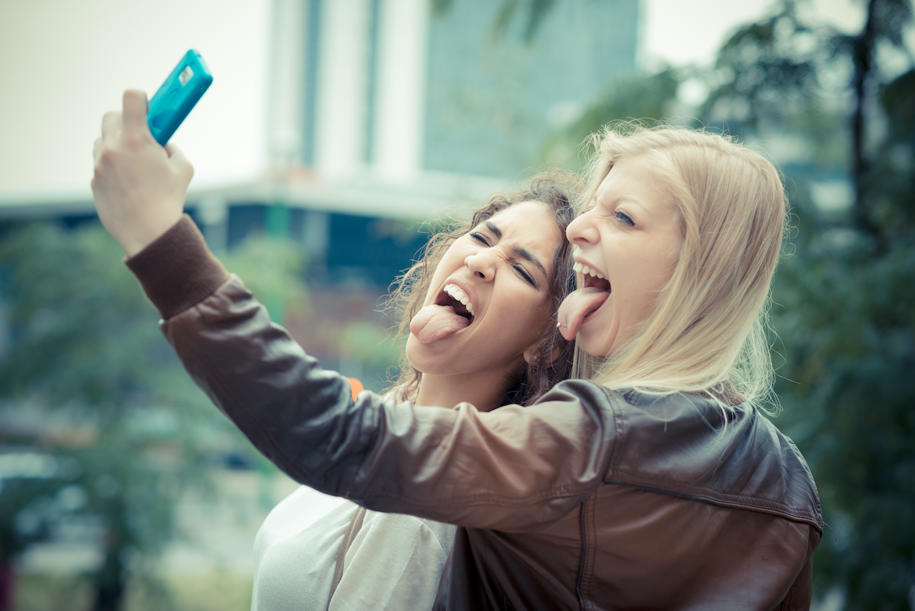 Na zdjęciu dwie zbuntowane nastolatki (z wypiętymi językami) robią sobie telefonem selfie.