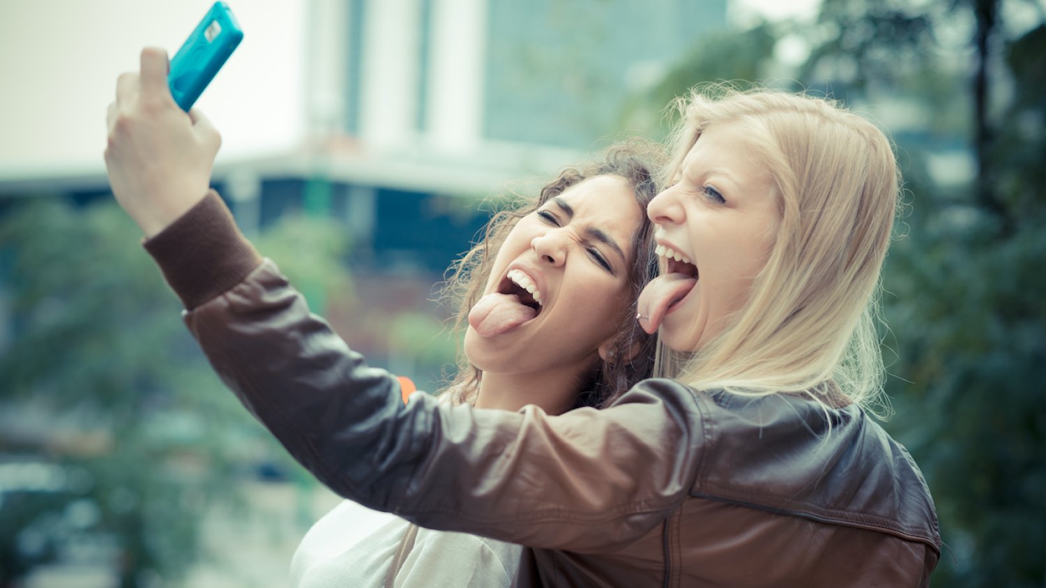 Na zdjęciu dwie zbuntowane nastolatki (z wypiętymi językami) robią sobie telefonem selfie.