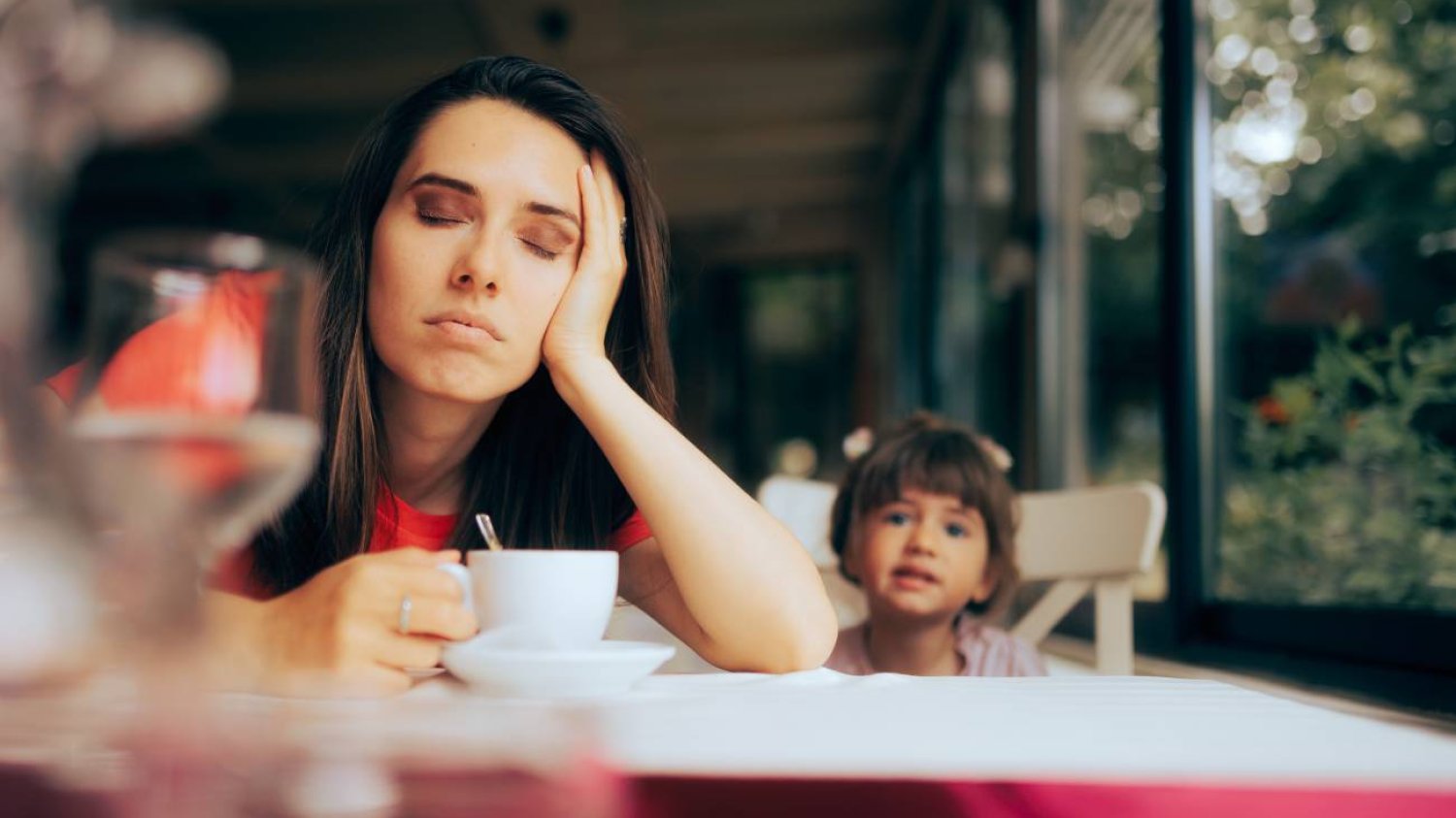 Mama zmęczona po pracy przysypia nad kawą, obok siedzi dziecko.