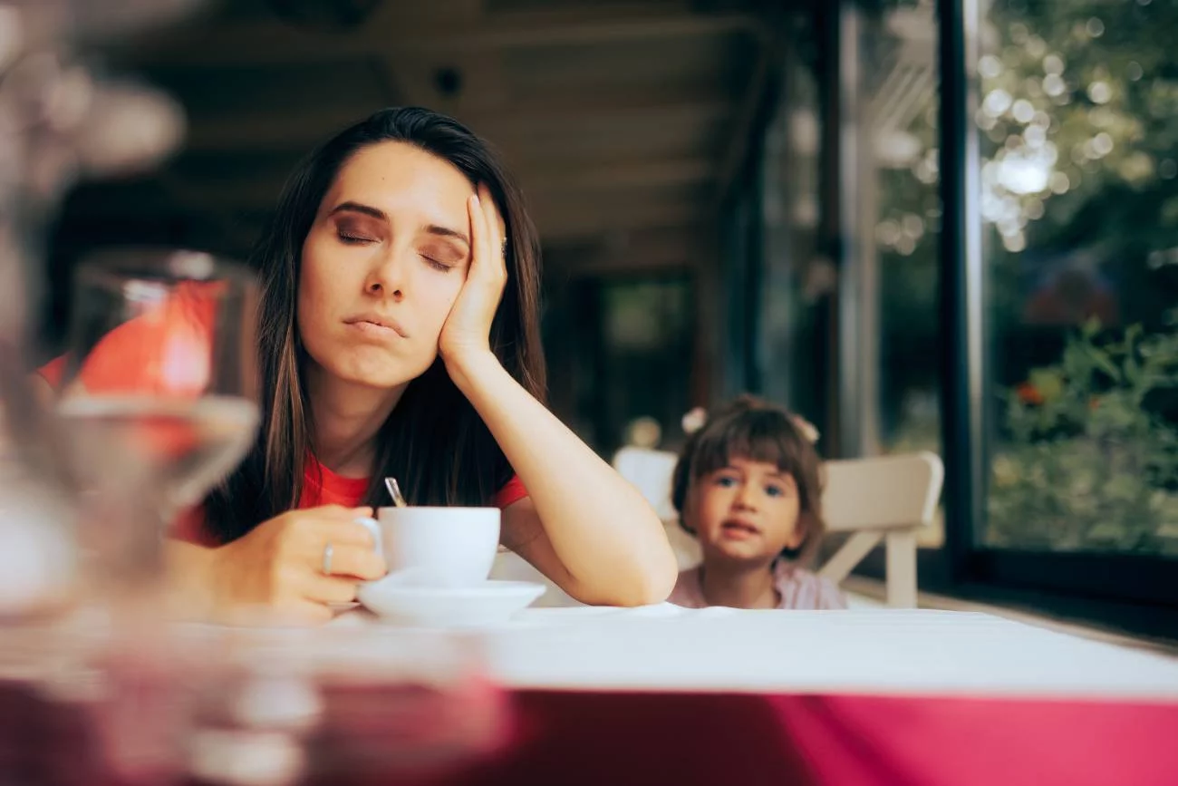 Mama zmęczona po pracy przysypia nad kawą, obok siedzi dziecko.