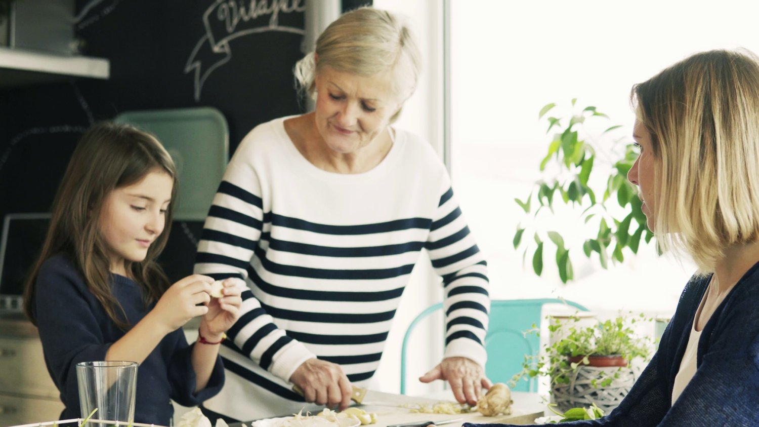 Na zdjęciu babcia z wnuczką przygotowują posiłek. Wszystkiemu przygląda się mama dziewczynki.