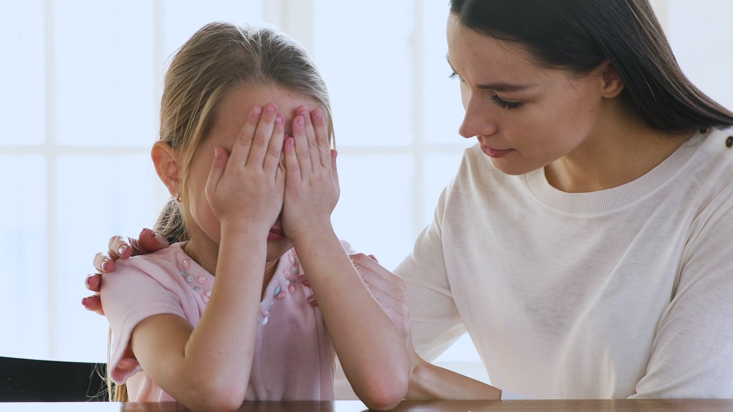 Dziecko pyta, czy umrze – jak powinno się odpowiedzieć? Na zdjęciu mała dziewczynka siedzi zasłaniając rączkami twarz, obok siedzi jej mama, próbując wytłumaczyć coś małej.