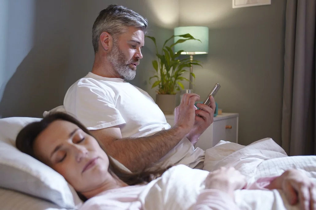 Na zdjęciu widać małżeństwo w jednym łóżku, ale każde z małżonków zajmuje się swoimi sprawami