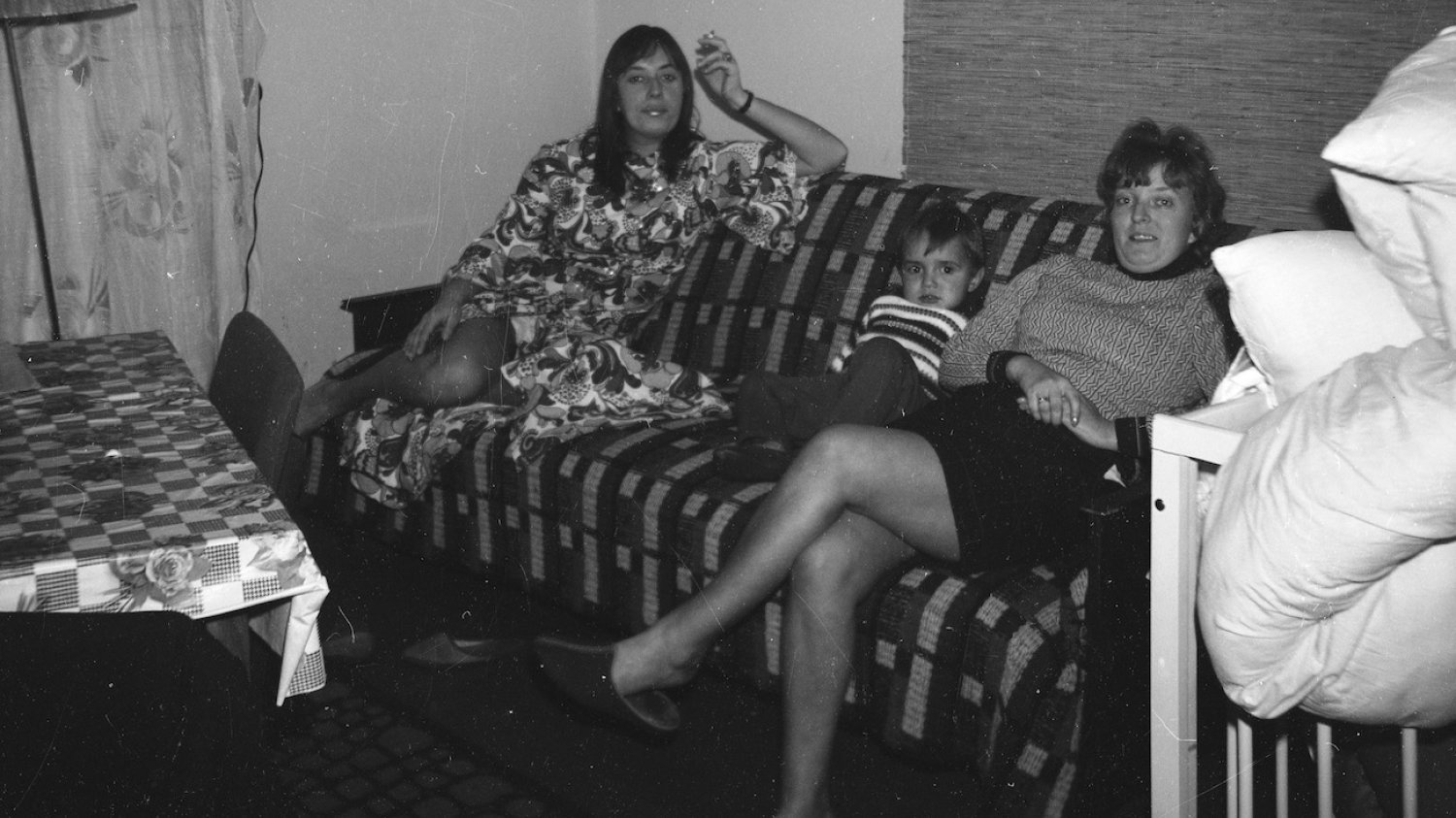Wnętrze mieszkania w 1971, na łóżku siedzą dwie kobiety z dzieckiem