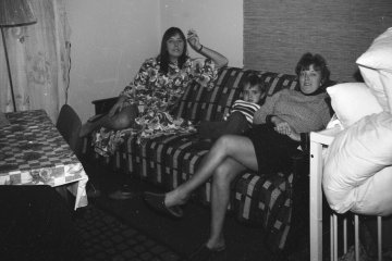 Wnętrze mieszkania w 1971, na łóżku siedzą dwie kobiety z dzieckiem
