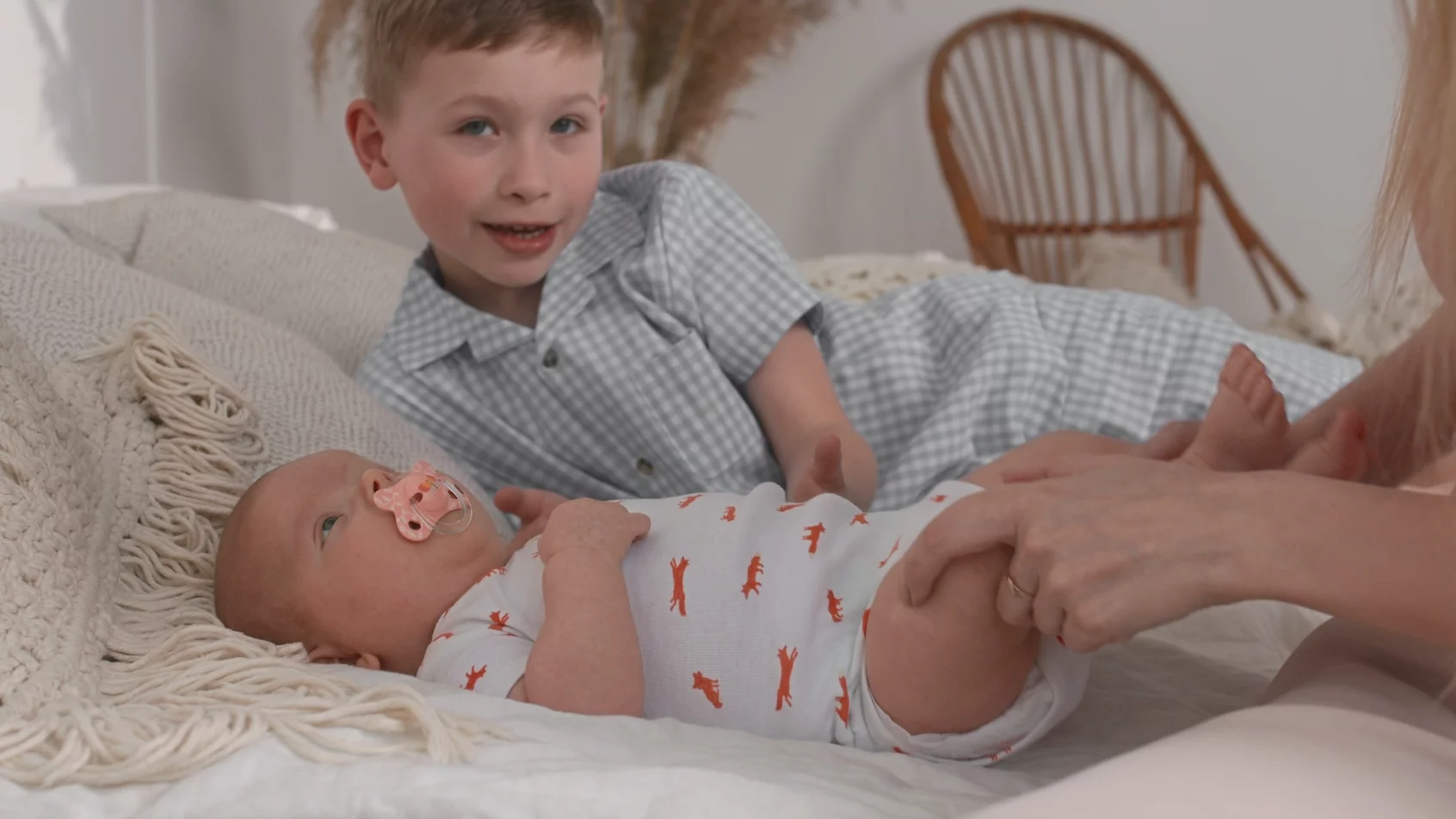 Jak reagować, kiedy dziecko jest zazdrosne o nowonarodzone rodzeństwo? Na zdjęciu kilkuletni chłopiec leży na łóżku z noworodkiem – widać też ręce mamy