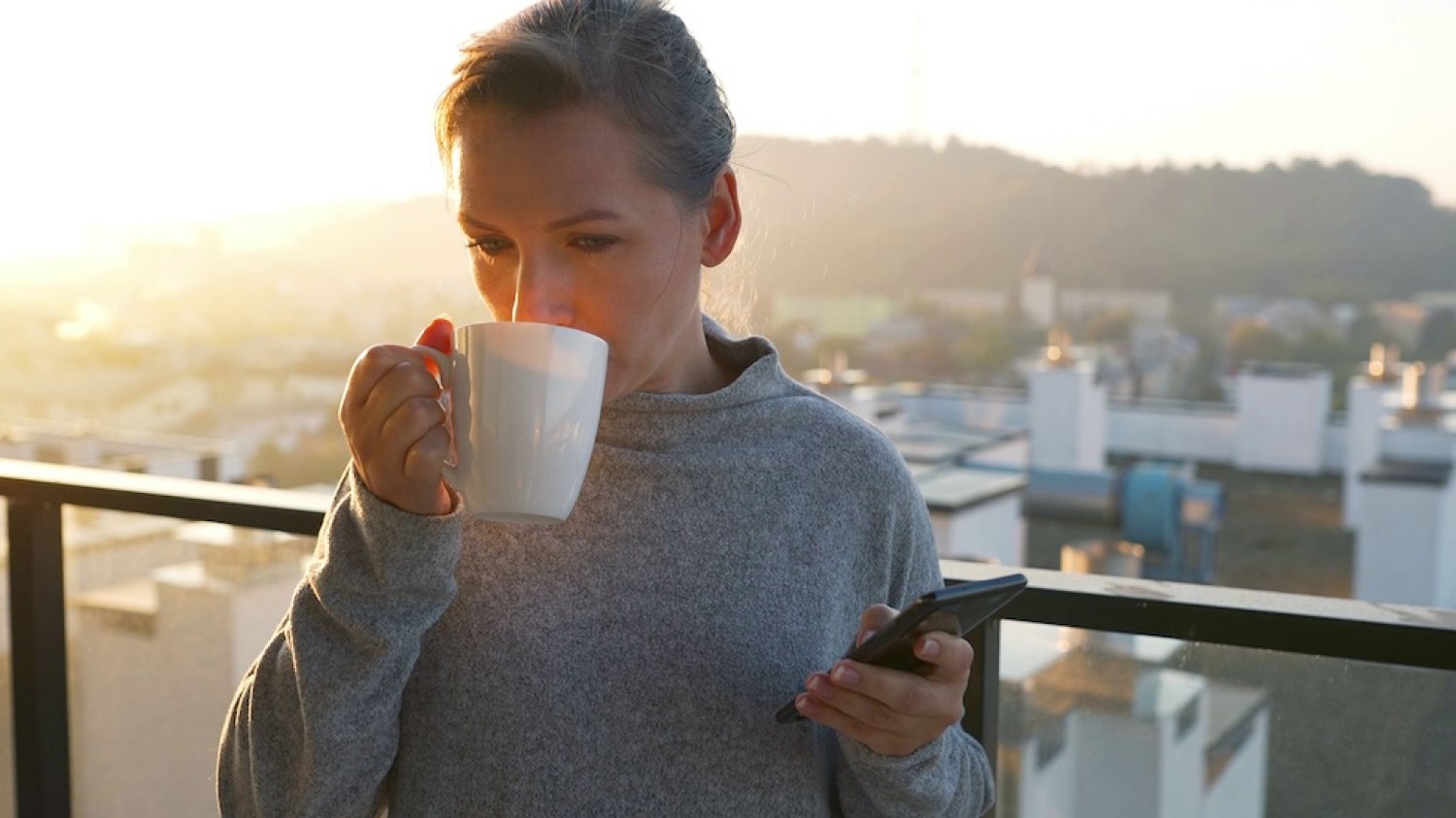 Na zdjęciu widać kobietę pijącą kawę i trzymającą w ręku telefon.
