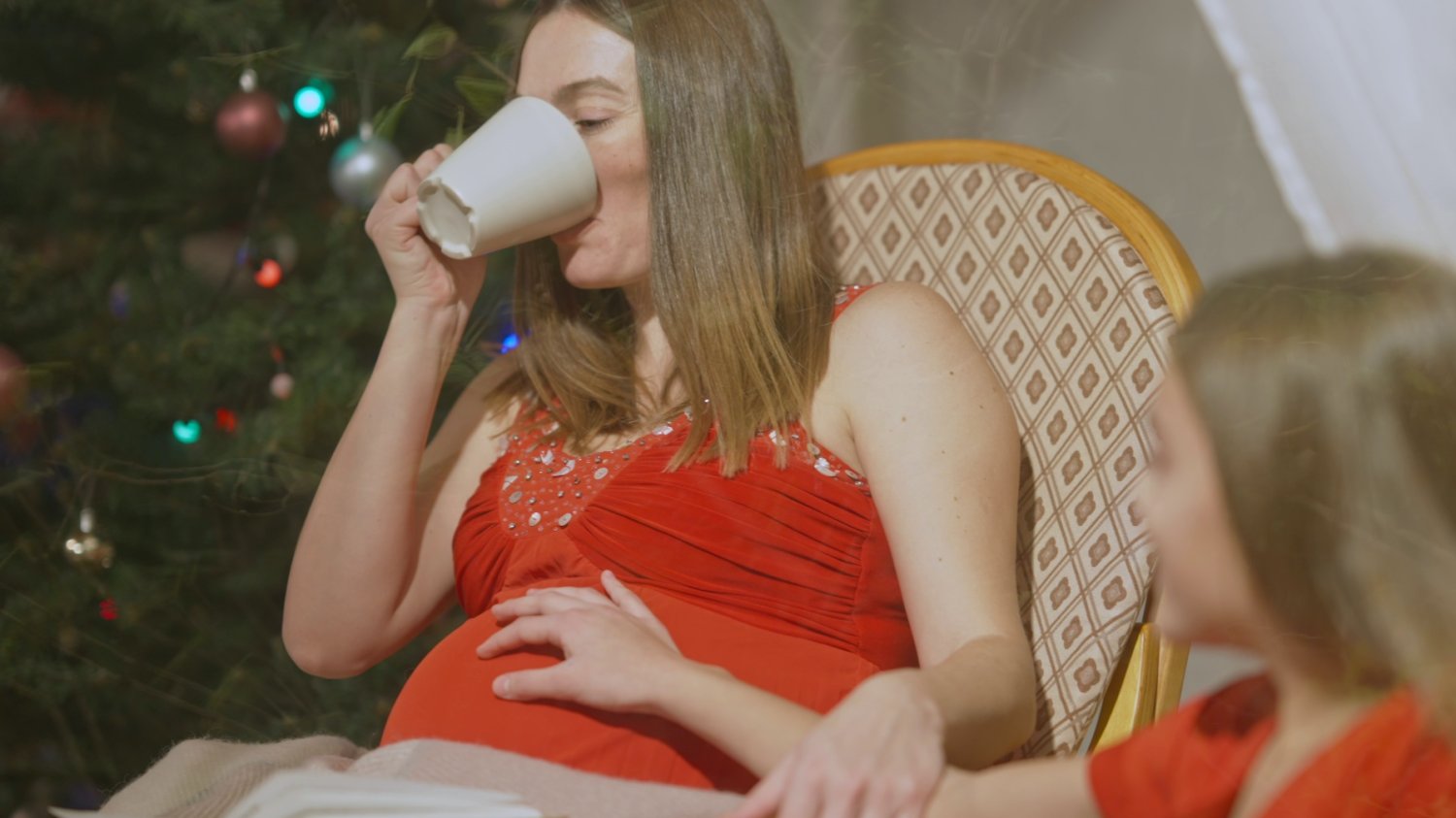 Na zdjęciu kobieta w ciąży siedzi przy choince i pije herbatę