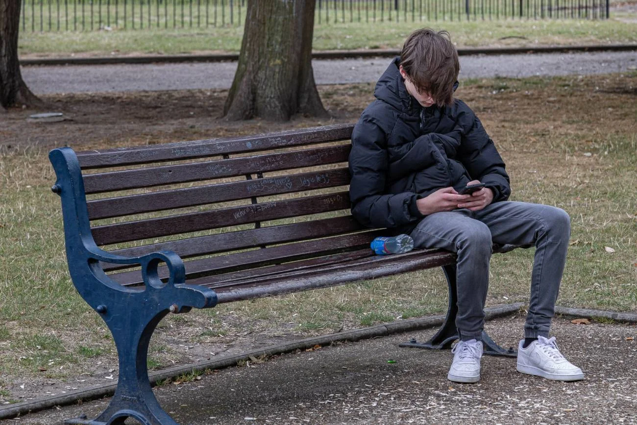 Nastolatek siedzi na ławce w parku ze smartfonem w dłoniach. Nastolatek siedzi na ławce w parku ze smartfonem w dłoniach.