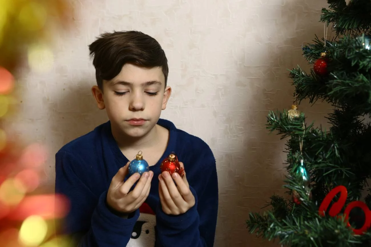 Smutny chłopiec ogląda bombki, siedząc obok choinki.