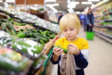 Mały chłopiec robi zakupy w markecie: wkłada ogórki do siatki.