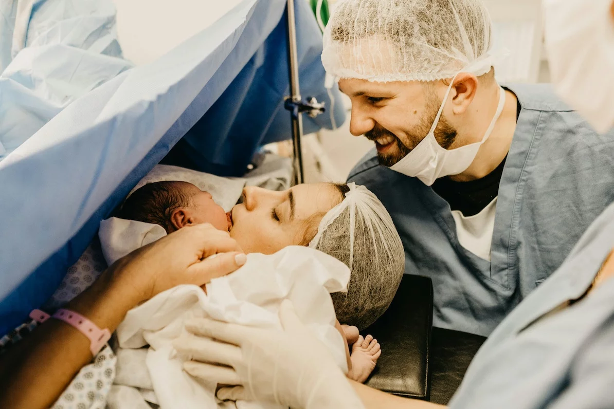 Na zdjęciu widać sytuację zaraz po porodzie - mama przytula noworodka. Tata stoi za nimi.