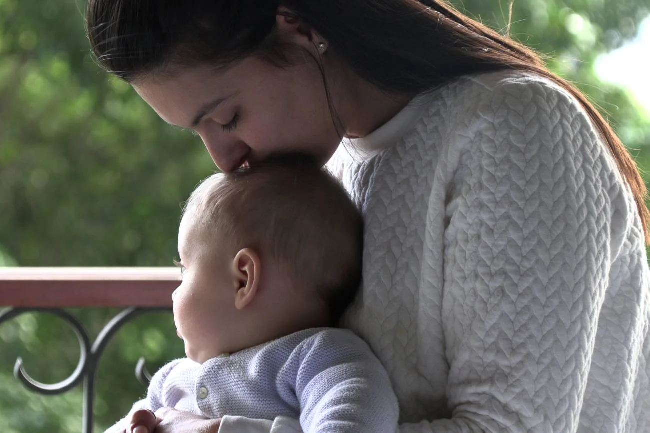 mama opiekuje się niemowlakiem, całuje go w główkę mama opiekuje się niemowlakiem, całuje go w główkę