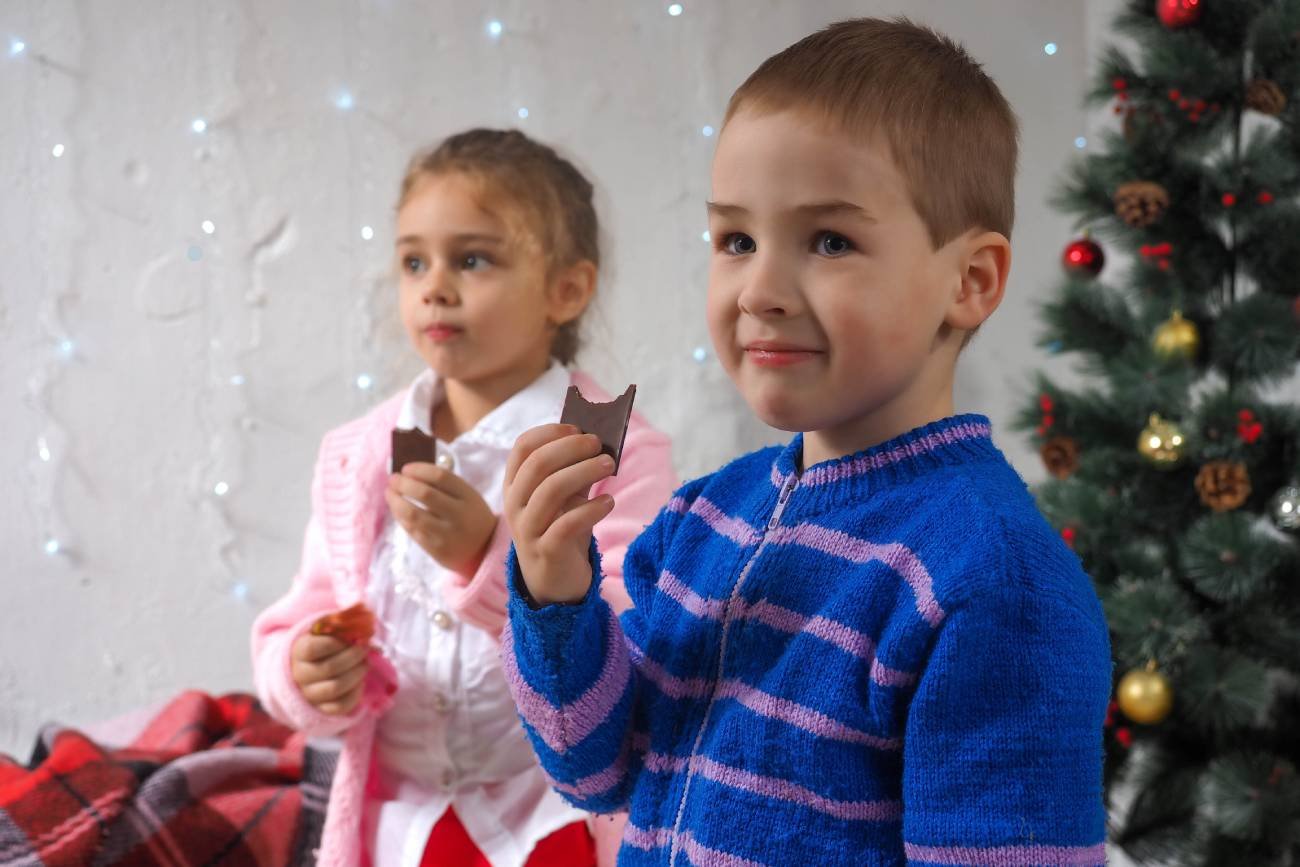 Dzieci jedzą czekoladę obok choinki.