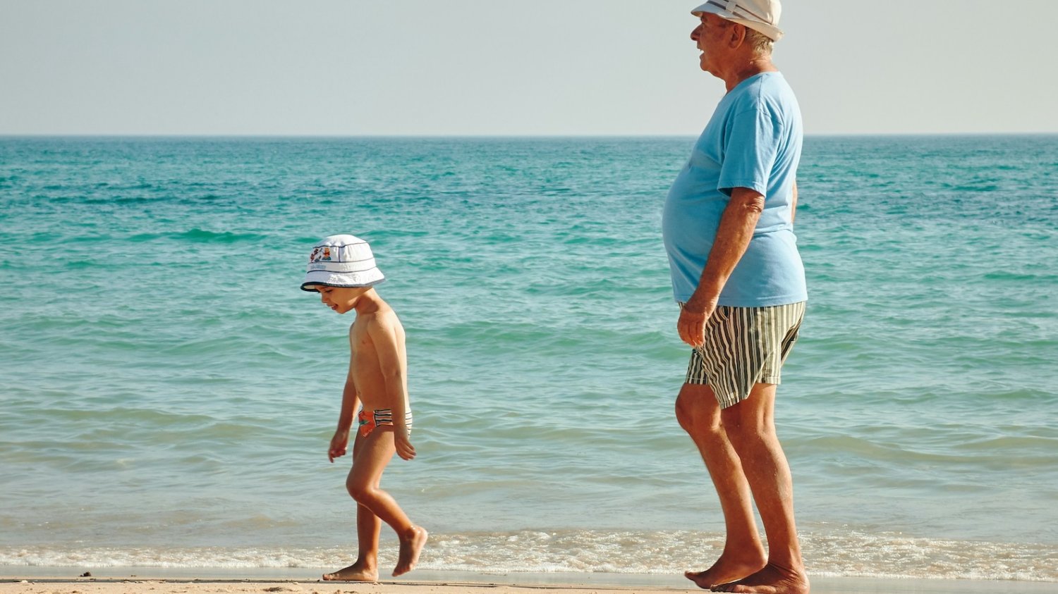 Na zdjęciu dziecko spaceruje z dziadkiem po plaży.