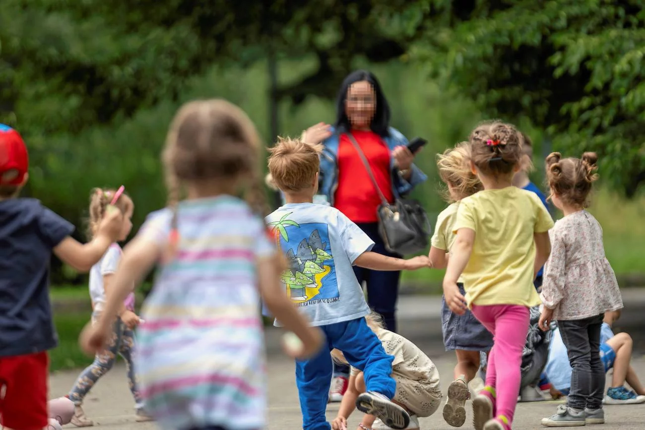 Dzieci z przedszkola na placu zabaw. Biegną do kobiety, która zajmuje się nimi, ale nie jest nauczycielką